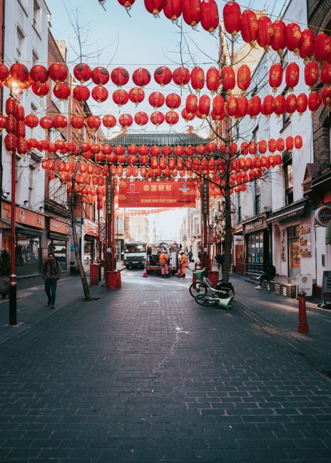 A street decorated with multiple rows of vibrant red Chinese lanterns hanging overhead, creating a festive atmosphere in Chinatown. The street is lined with shops and eateries, with a few pedestrians 