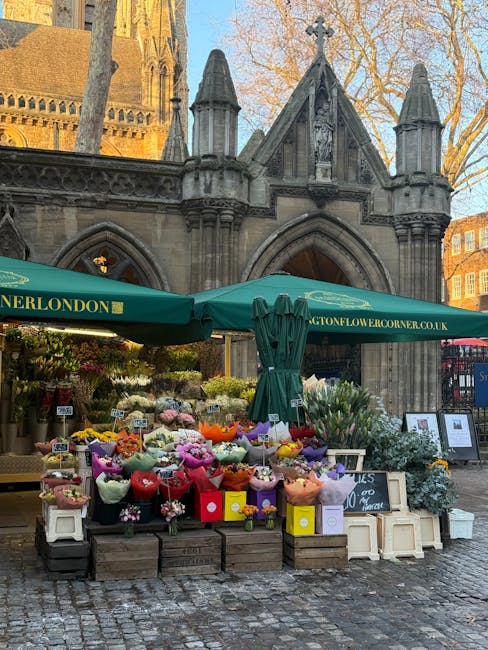 A vibrant outdoor flower stall situated in front of a historic stone building with gothic architectural details, including pointed arches and decorative pinnacles. The stall is shaded by a green canop