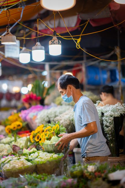 A florist wearing a light blue face mask and casual clothing inspects a bouquet of white chrysanthemums at a bustling flower stall in a market. The stall is filled with an array of vibrant flowers inc