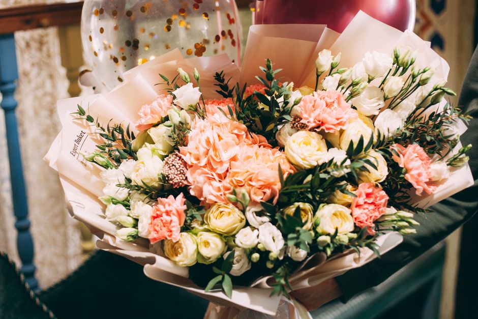 A floral bouquet held by a person, featuring a mix of fresh flowers including large peach-colored carnations, creamy white roses, and smaller white lisianthus, accented with dark green foliage. The ar