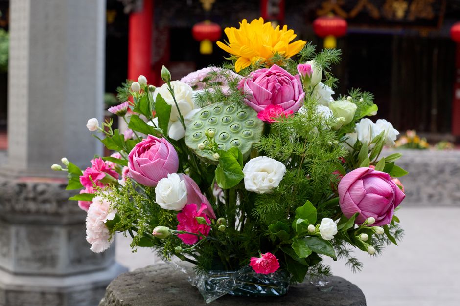 A floral arrangement featuring pink lotus flowers, white lisianthus, and small pink carnations, complemented by green foliage and sprigs of ferns. The bouquet is placed in a clear glass vase, with vis