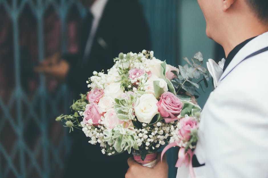 A man dressed in a white tuxedo with black trim holds a floral bouquet composed of pink and white roses, white baby's breath, and sprigs of dusty miller foliage, wrapped in pink wrapping paper with a